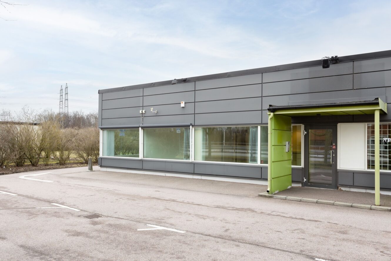 En modern, envånings kommersiell byggnad med grå panel, stora fönster och ett grönt entrétak, belägen intill en asfalterad parkeringsplats.