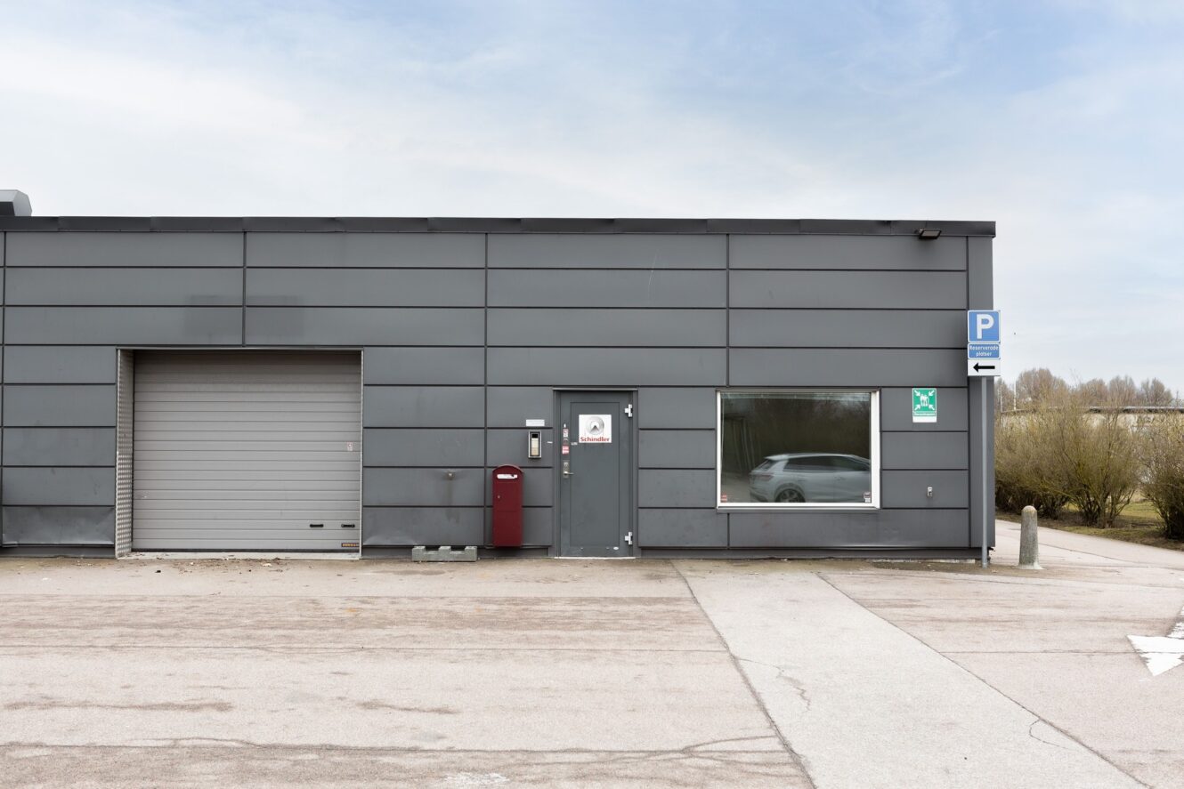 En grå industribyggnad med en stängd garageport, en röd brevlåda, en glasdörr, ett stort fönster och en parkeringsskylt; en bil speglar sig i fönstret.