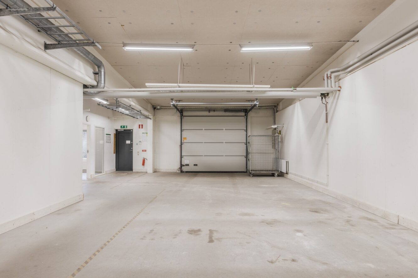 Tomt industrigarage med vita väggar, betonggolv, lysrörsbelysning och en stängd takskjutport längst bort. Ledningsrör och elpaneler är synliga.