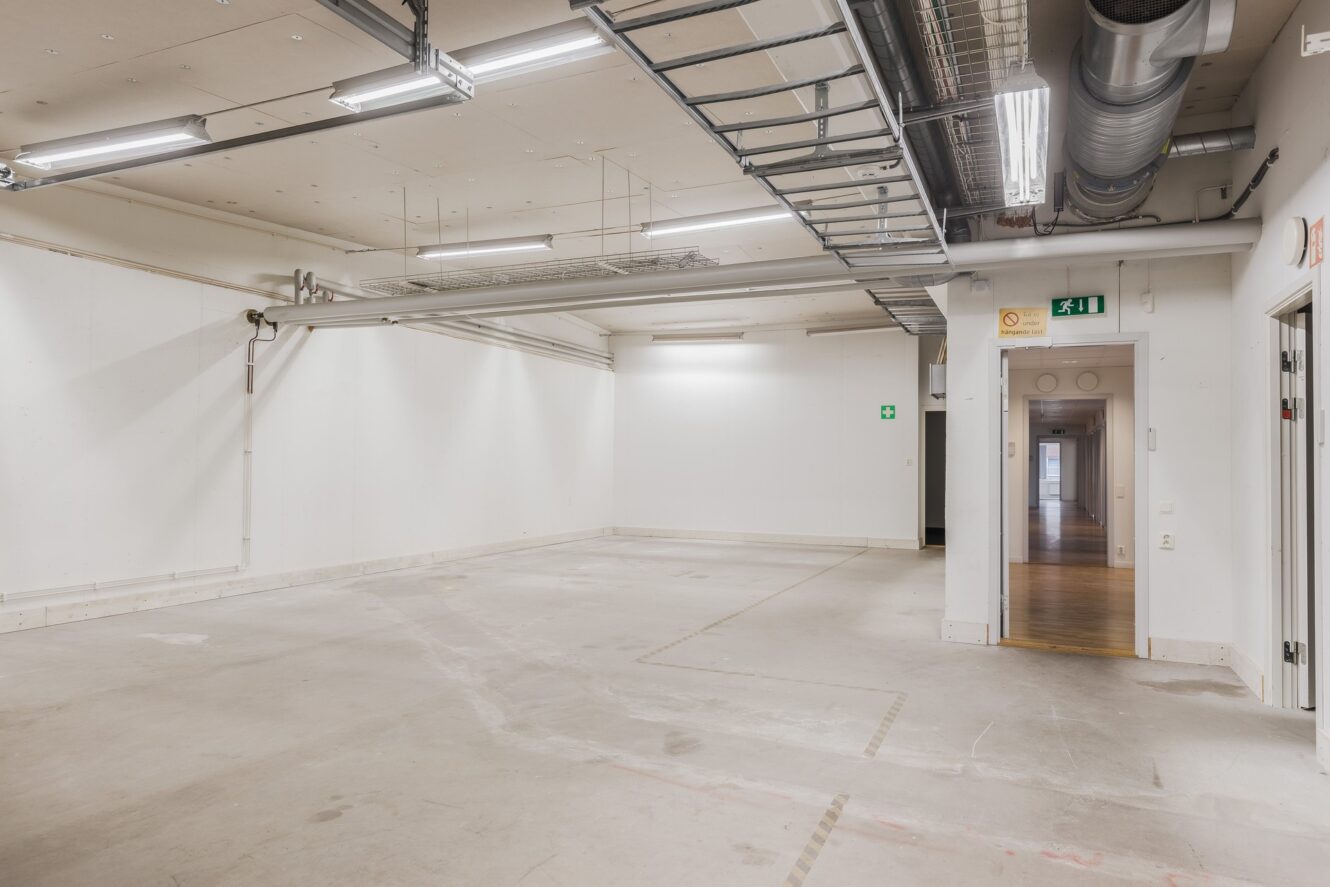 Tomt industrirum med synliga rör och kablar i taket, vita väggar, betonggolv och en dörröppning som leder till en hall med trägolv.