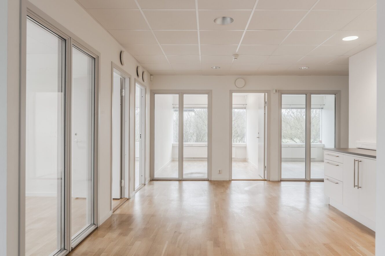Ett ljust, tomt kontorsutrymme med glasdörrar, stora fönster, ljust trägolv och vita väggar, sett från en öppen yta nära ett litet pentry.