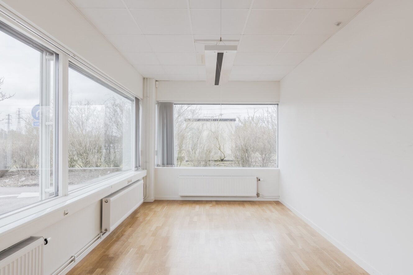 Tomt, omöblerat rum med vita väggar, stora fönster på två sidor, trägolv och en taklampa. Naturligt ljus kommer in genom fönstren och visar en utsikt över träd utanför.