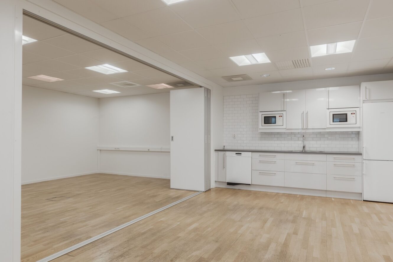 Ett modernt, tomt rum med trägolv och vita väggar, anslutet till ett kök med vita skåp, två mikrovågsugnar och ett kylskåp.