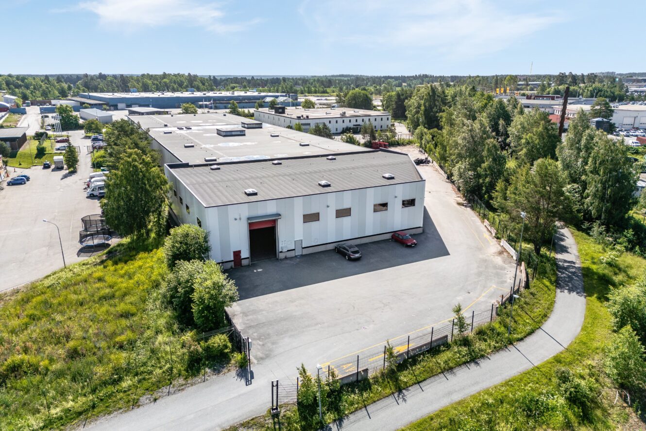 En stor industrihall med vit fasad och lastkajer, omgiven av grönska och en asfalterad väg, belägen i ett företagsområde.