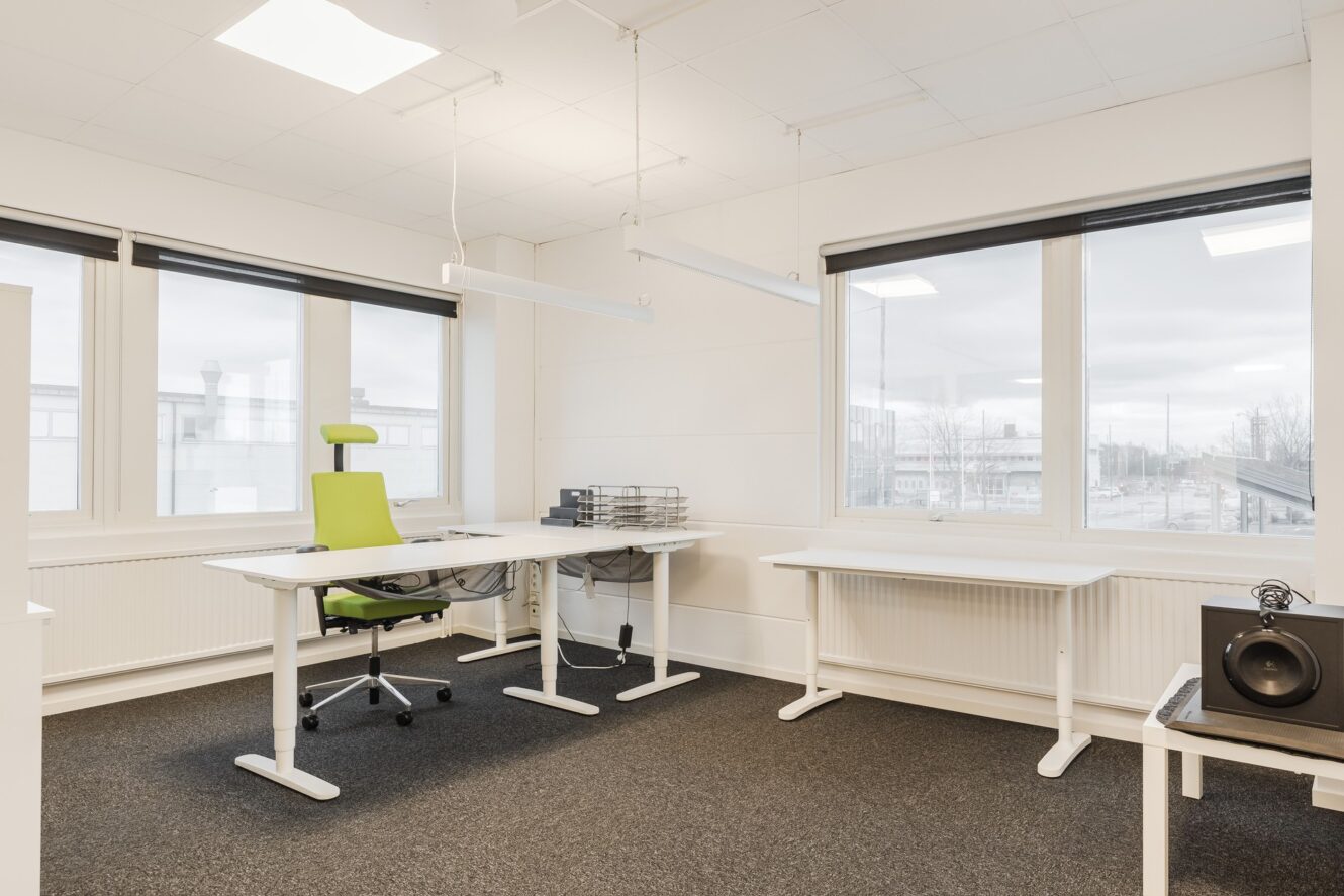 Modernt kontor med vita väggar, stora fönster, två skrivbord, en grön kontorsstol, dokumenthyllor och en högtalare på ett sidobord. Utrymmet är ljust och minimalt möblerat.