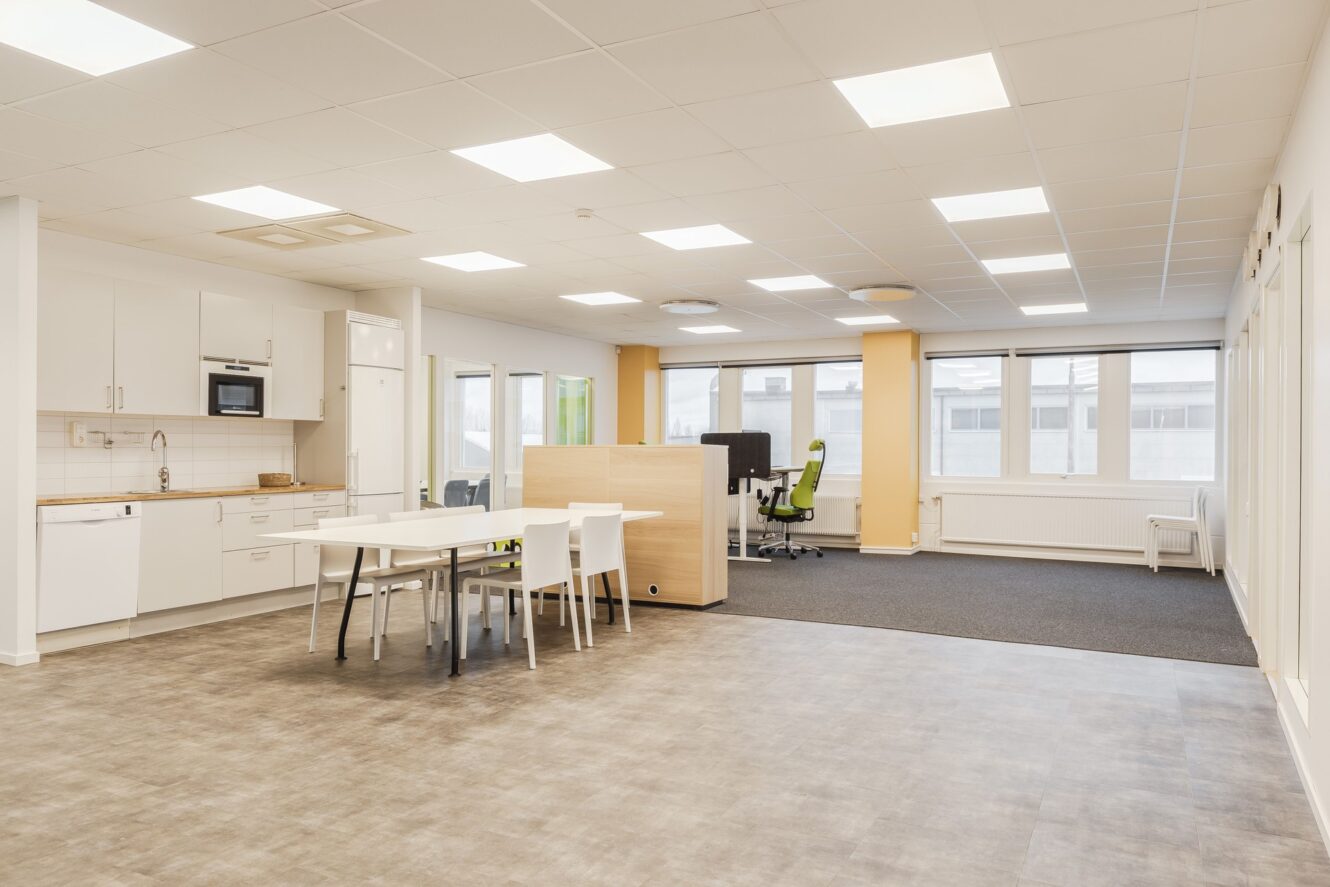 Moderna kontorslokaler med pentry, matbord med stolar, öppet arbetsområde med skrivbord och stolar, stora fönster och stark takbelysning.