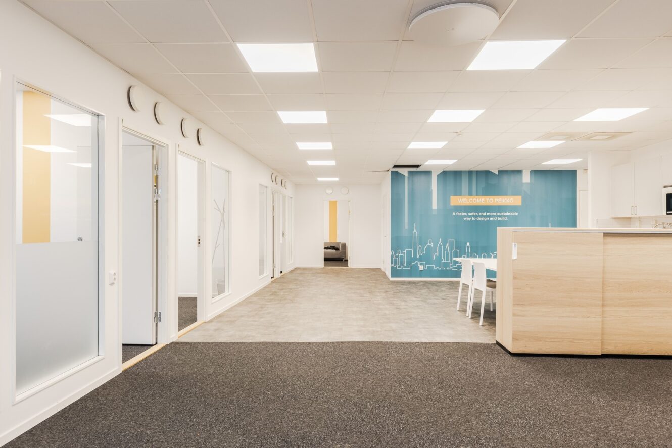 Modern kontorsinredning med vita väggar, stark belysning, ett pentry till höger, flera dörrar till vänster och en blå accentvägg med ett välkomstmeddelande i bakgrunden.