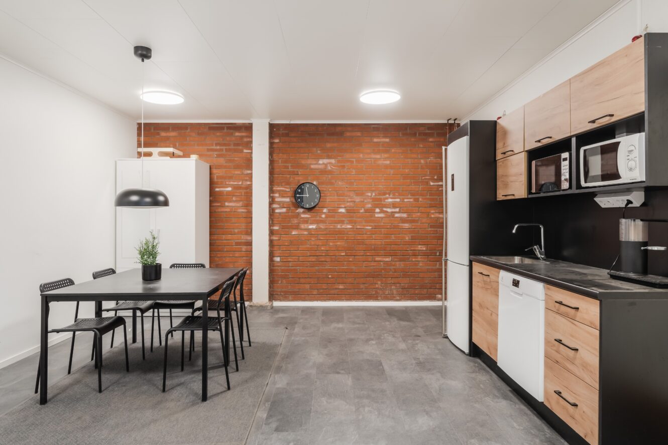 Modernt kök och matplats med svart matbord och stolar, skåp med träaccenter, vita vitvaror och en accentvägg i tegel med en rund klocka.