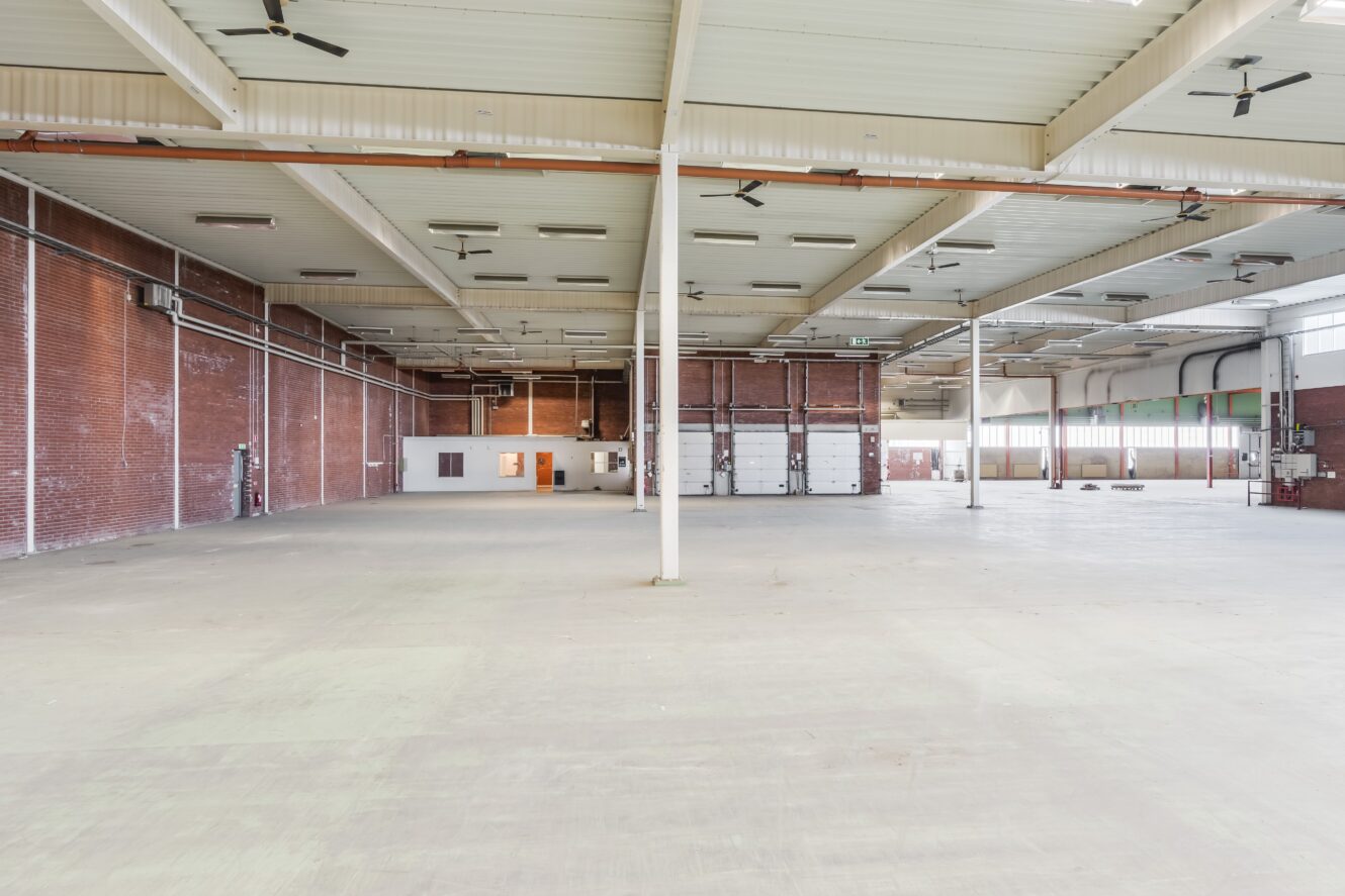 Stor, tom industrilokal med högt i tak, synliga bjälkar, tegelväggar och ett litet kontorsutrymme på baksidan.