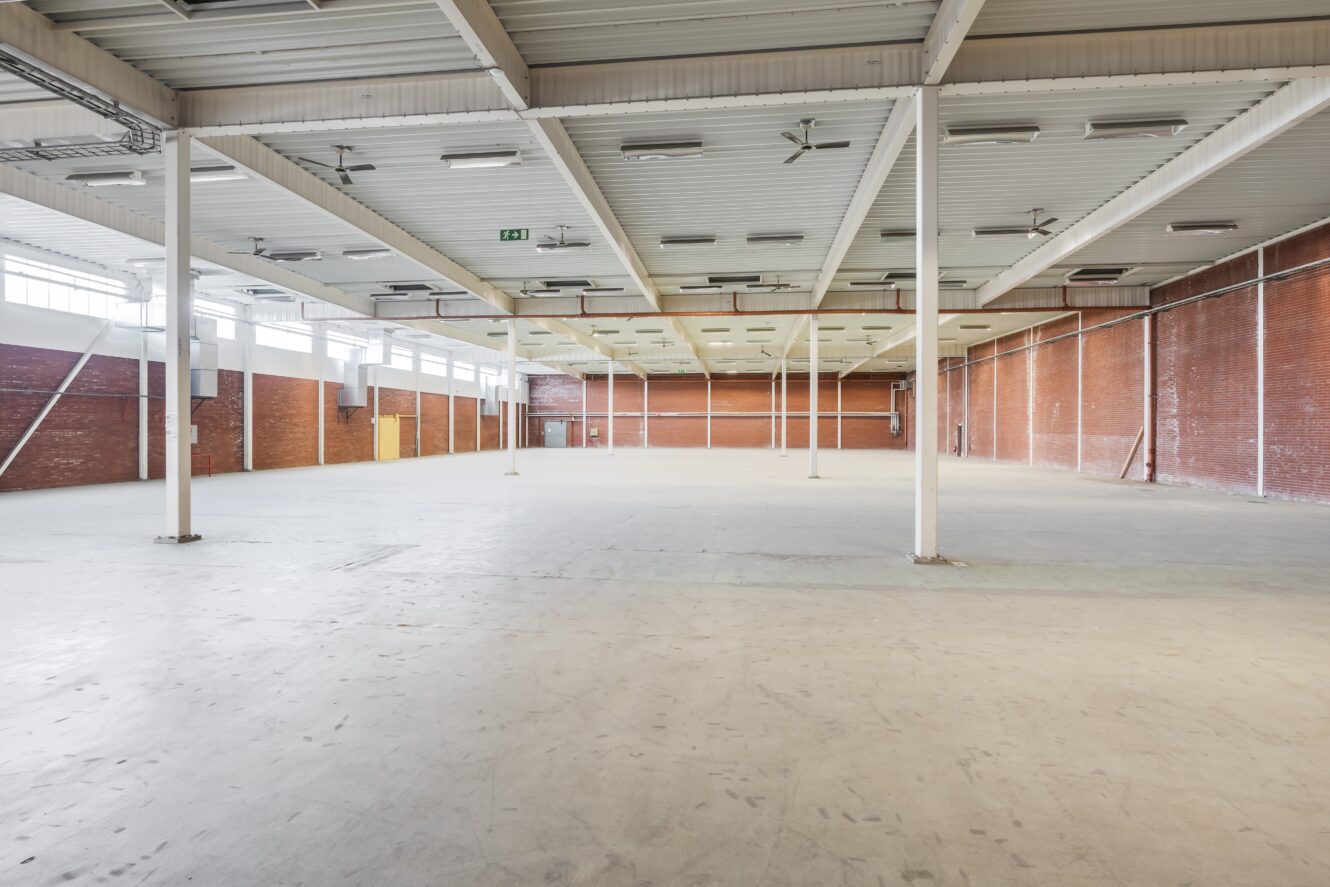 Stor tom lagerlokal med betonggolv, tegelväggar, metalltak och stödpelare. Taklampor och fönster längs de övre väggarna ger belysning.