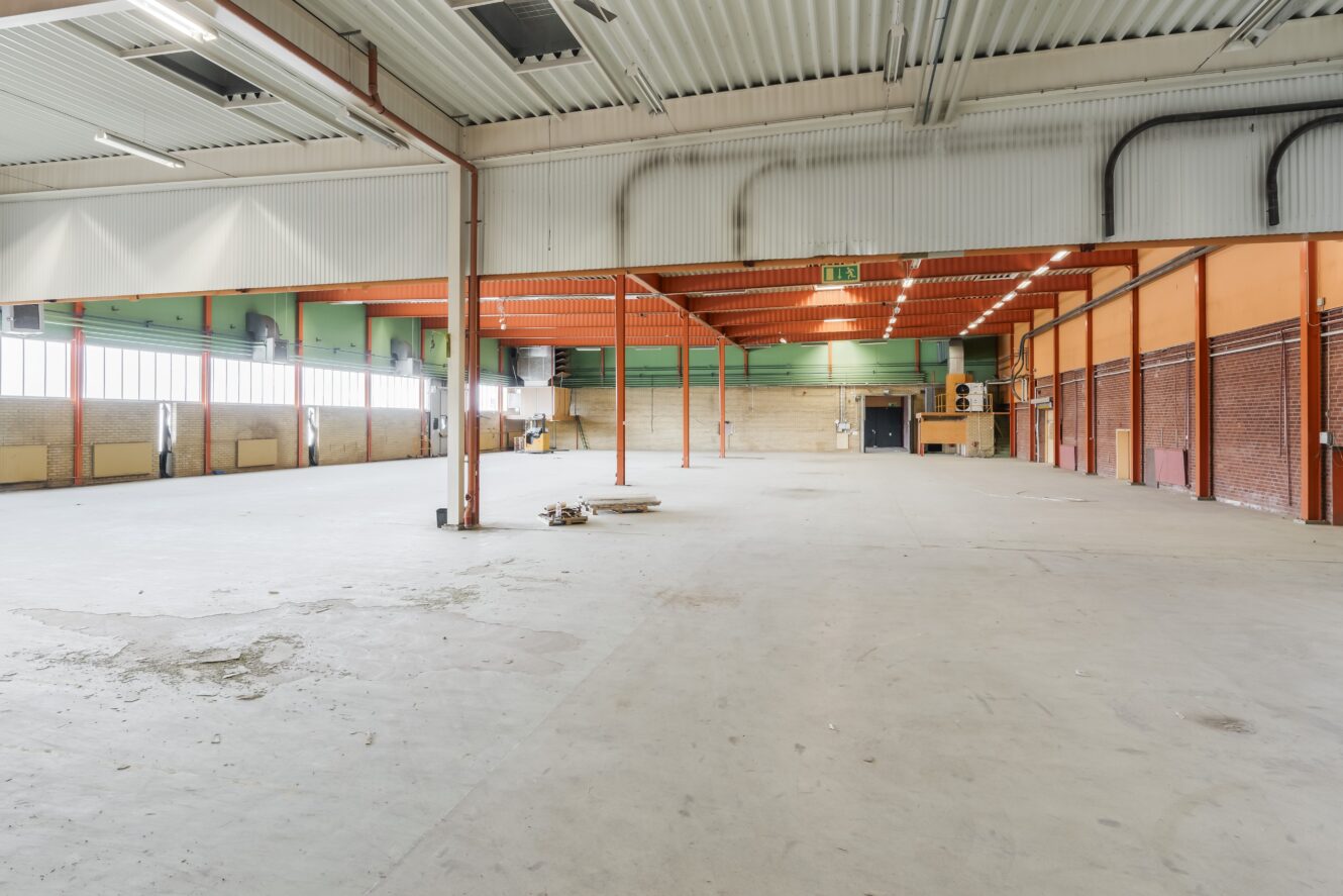 Stor, tom industrilokal med högt i tak, synliga bjälkar och betonggolv. Naturligt ljus kommer in genom fönster längs ena väggen. På golvet syns en del utspritt bråte.