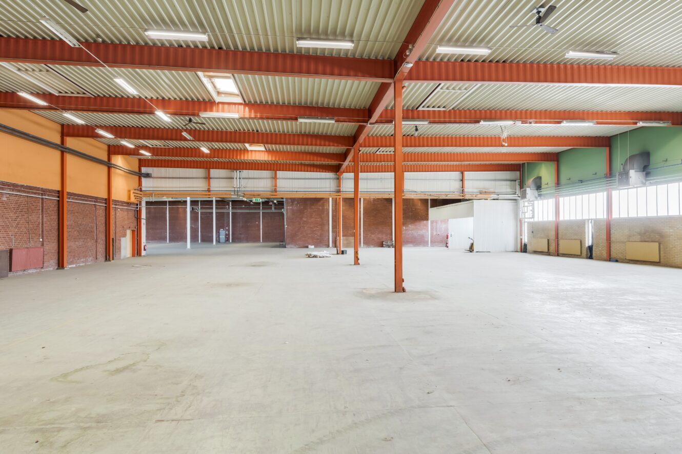 Rymlig, tom lagerlokal med högt i tak, synliga bjälkar, tegelväggar, stora fönster och betonggolv. En del bråte och byggmaterial utspridda på golvet.