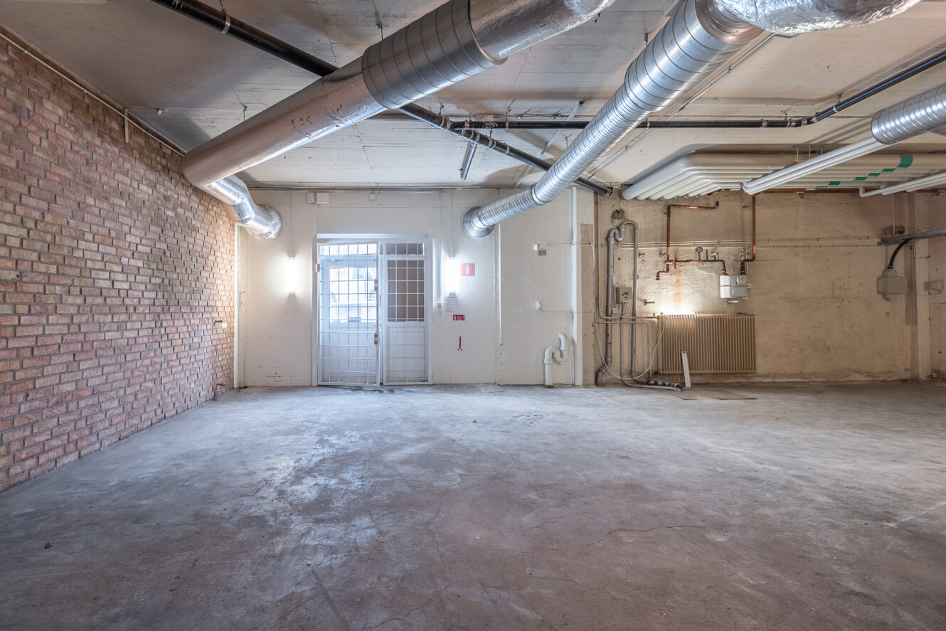 Tomt industriellt rum med exponerad tegelvägg, synligt kanalsystem i taket, betonggolv och en metalldörr med glaspaneler som släpper in naturligt ljus.