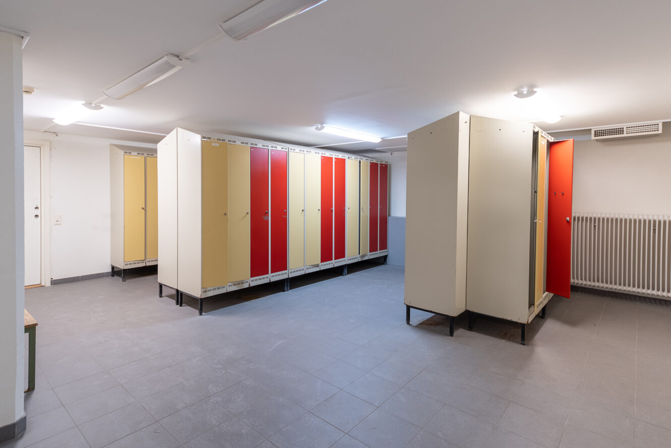 Ett rent omklädningsrum med rader av beige, gula och röda skåp, klinkergolv, taklampor och ett element mot väggen.