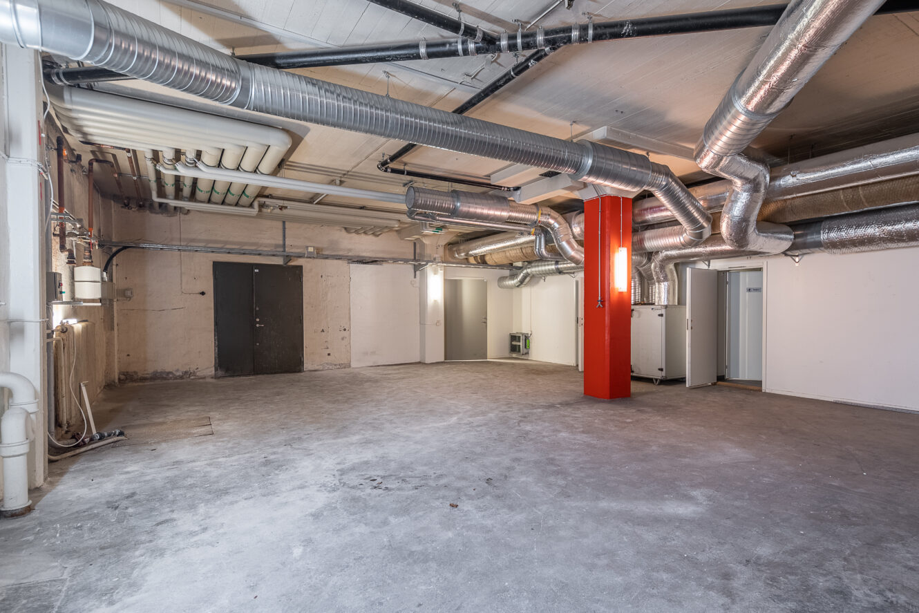 Stor, tom källare med synliga rör och kanalsystem i taket, betonggolv, vita och beige väggar och en framträdande röd pelare.