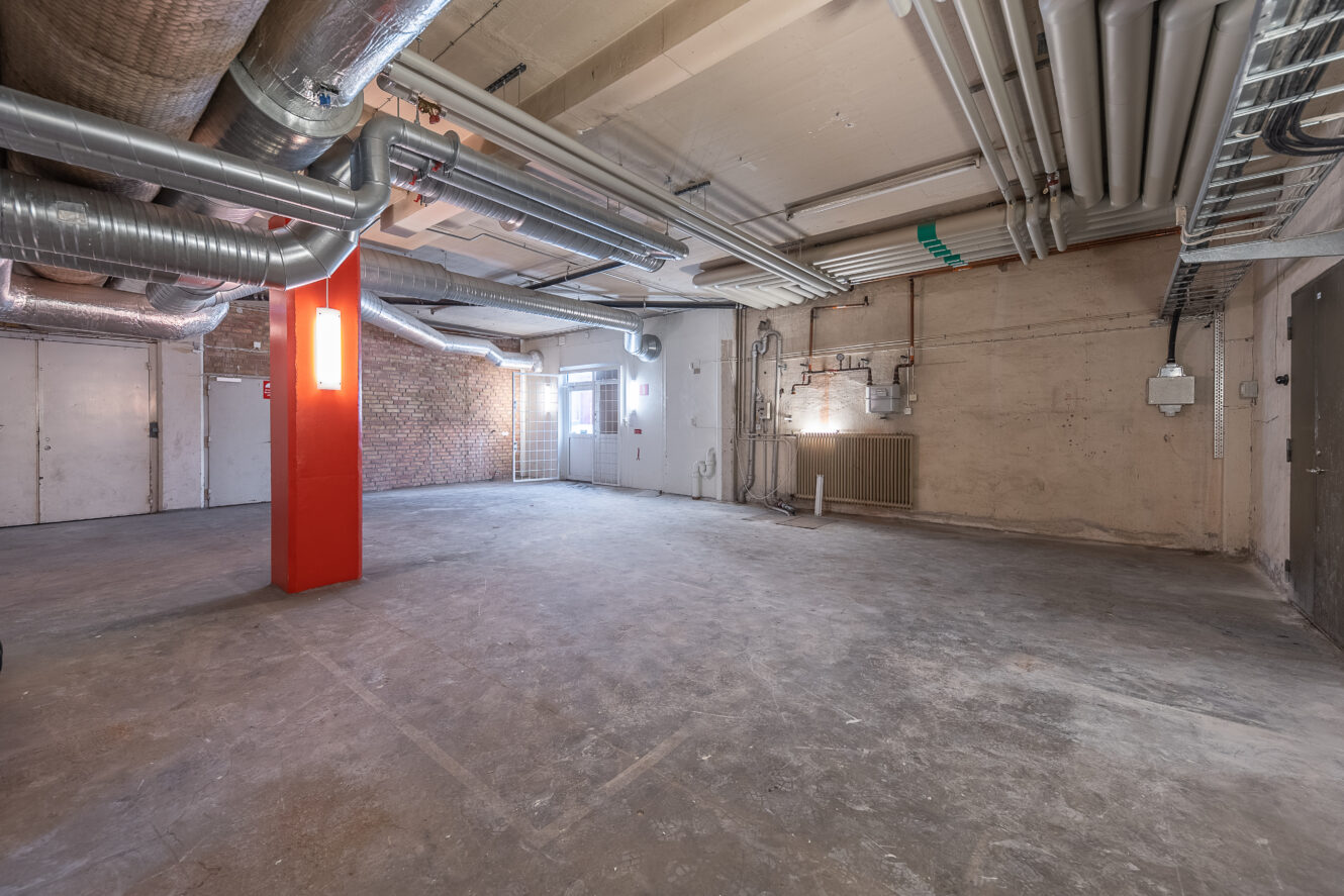 Ett tomt industrirum med synliga rör, kanaler och ledningar i taket, en röd pelare, betonggolv och väggar av tegel och puts. Naturligt ljus kommer in genom fönster på ena sidan.