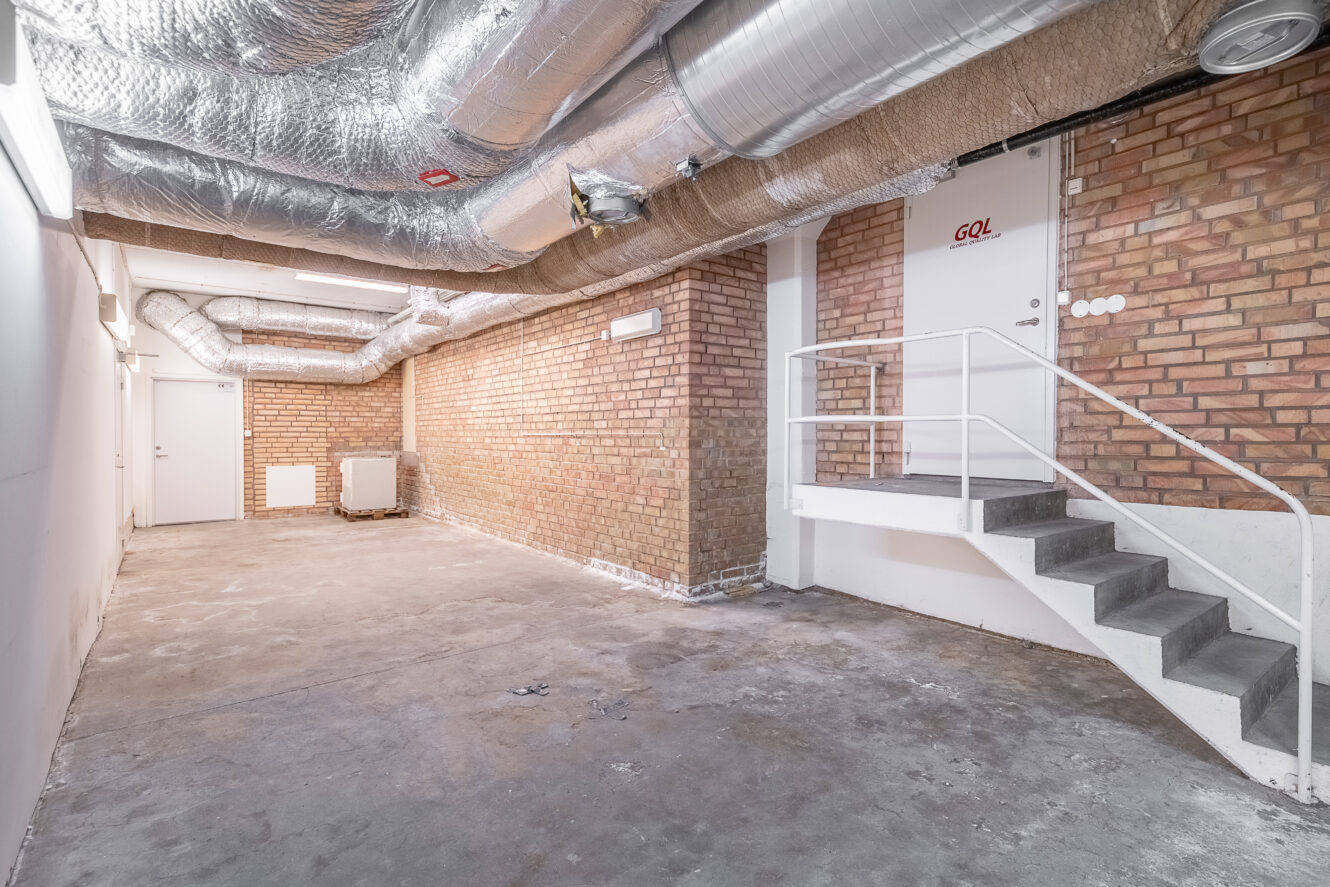 En källare med synliga tegelväggar, metalliska luftkanaler i taket, betonggolv, vita dörrar och en kort trappa som leder till en dörr märkt GOL.