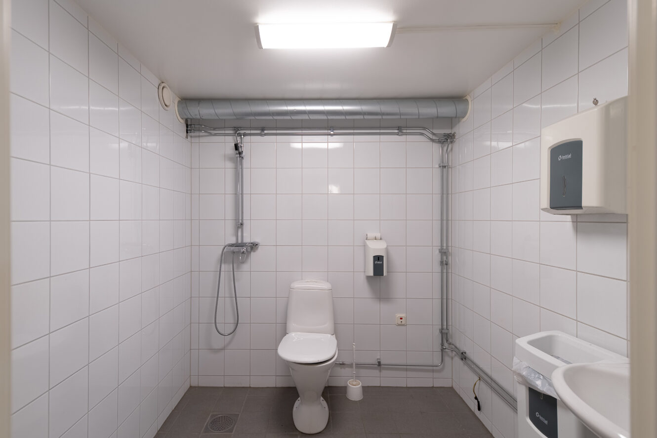 Ett toalettrum med toalett, väggmonterad dusch, toalettpappershållare, tvålpump och handfat. Väggarna är vitkaklade och det finns synliga rörledningar nära taket.