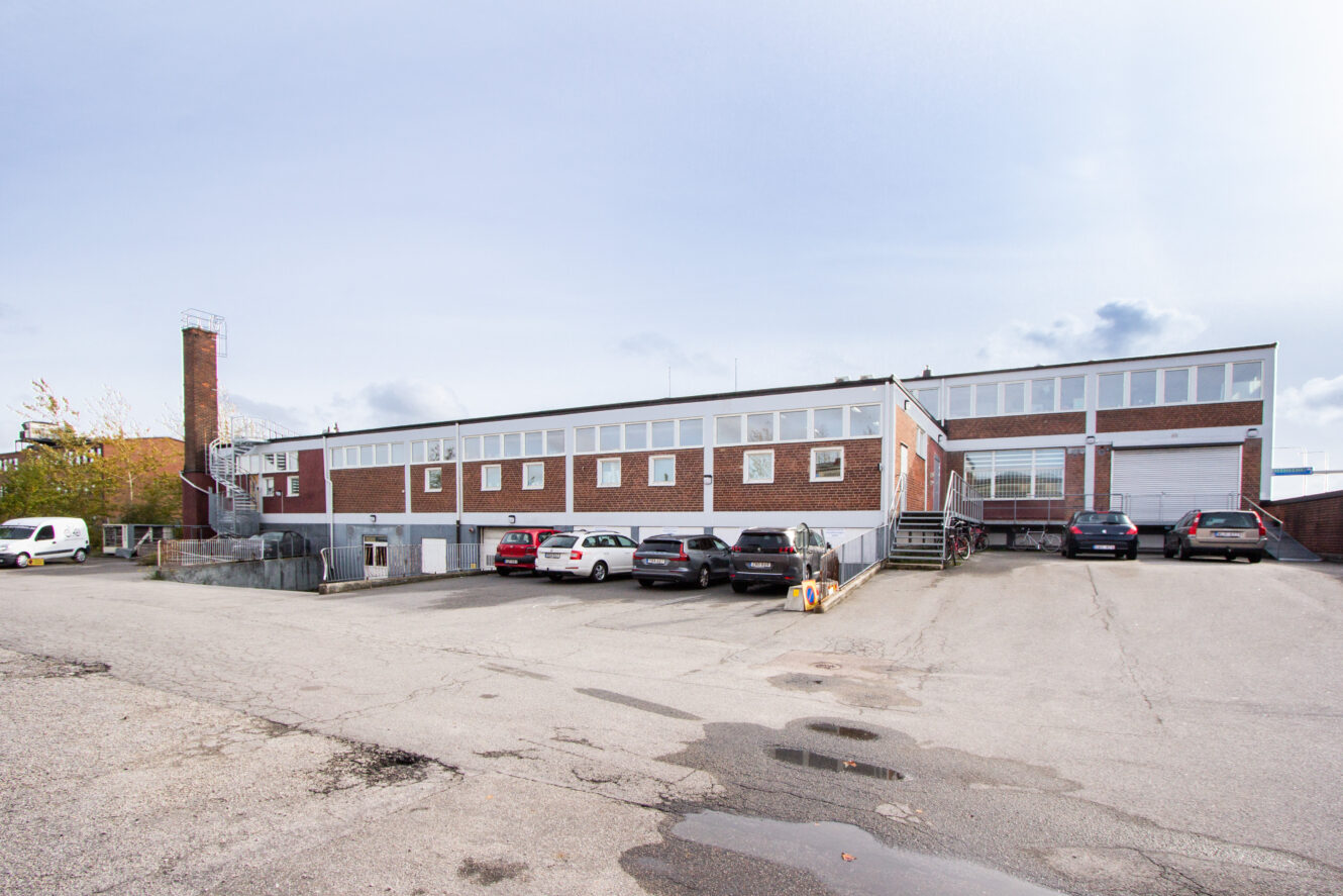 En tvåvånings tegelbyggnad med stora fönster, flera bilar parkerade framför och en metalltrappa på sidan på en asfalterad parkeringsplats.