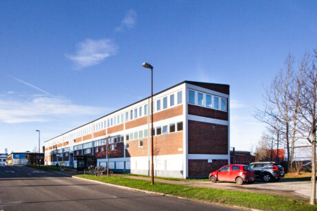 En trevåningsbyggnad i tegel och vitt med stora fönster, intill en gata med parkerade bilar, cyklar och en blå himmel ovanför.