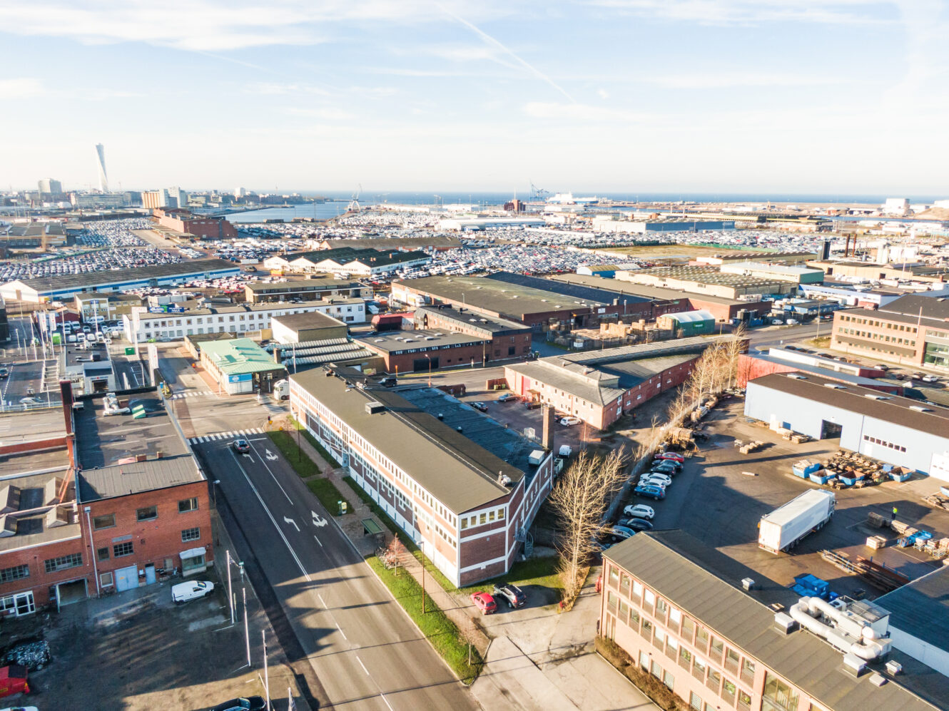 Flygfoto över ett industriområde med lagerbyggnader, parkeringsplatser och vägar nära en hamn, med havet och stadsbilden i bakgrunden.