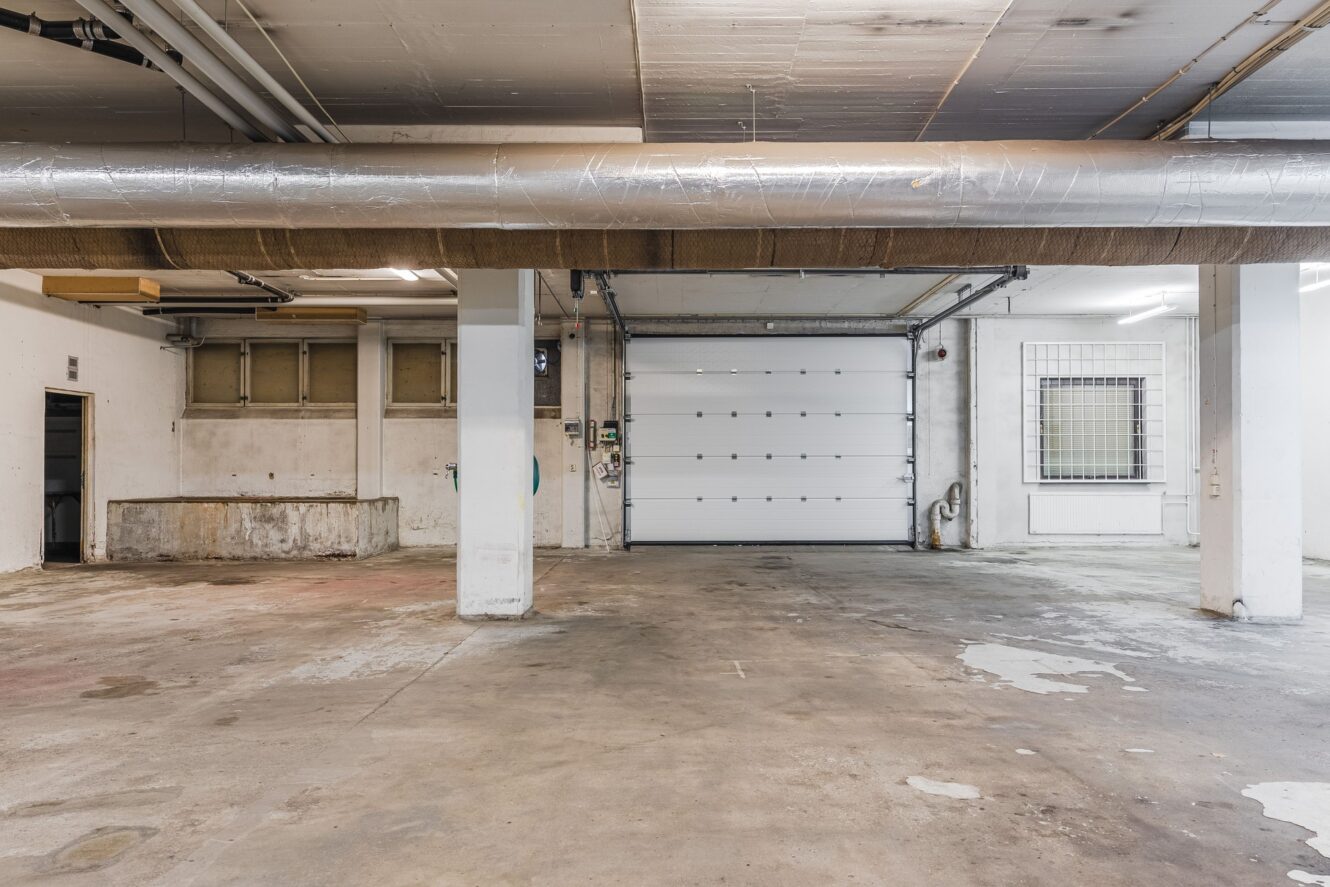 Tomt industrigarage med betonggolv, vita väggar, synliga rör och en stängd rullport. Ett fönster och elanslutningar syns på höger sida.