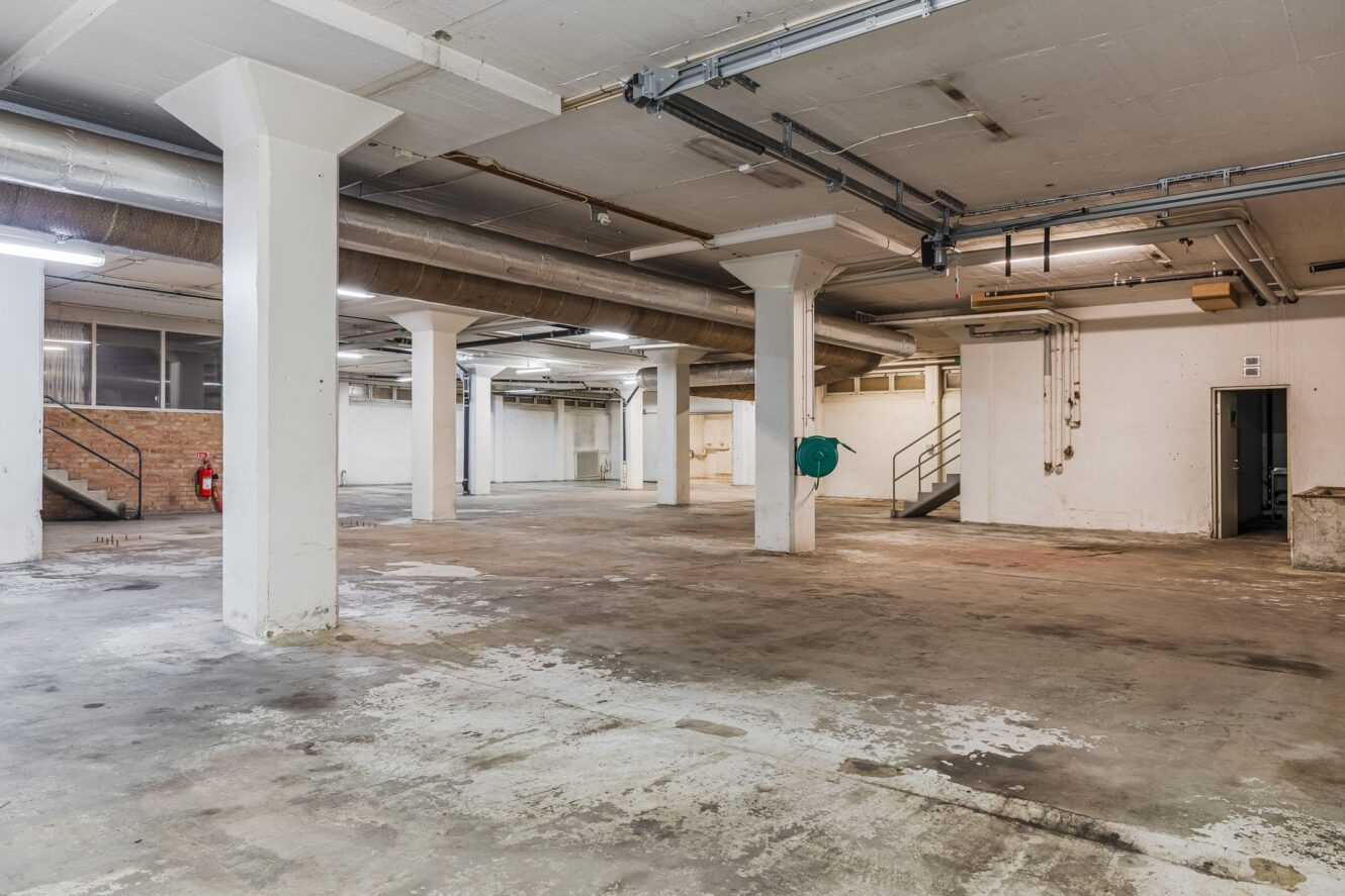 Stor tom industriell källare med betonggolv, tjocka pelare, synliga rör, tegelvägg, metalltrappa och verktygsutrustning på väggarna.