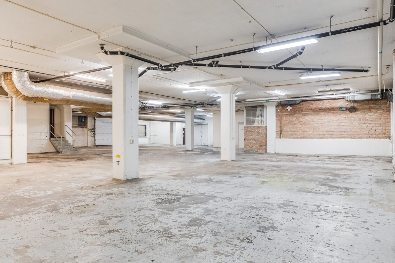 Rymlig, tom industrikällare med betonggolv, synliga rör, tegelväggar och vita väggar, stödpelare, lysrörsbelysning och en trappa som leder till en upphöjd yta.