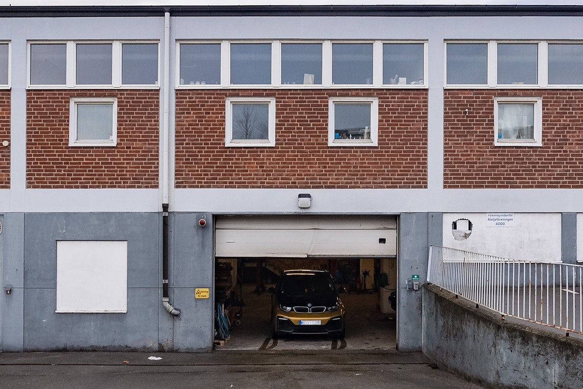 En svart bil står parkerad i ett garage med en delvis öppen dörr i en tvåvåningsbyggnad i tegel och betong.