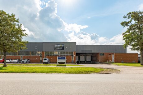 En stor industrihall med tegel och grå fasad, flera parkerade bilar, en skylt vid huvudentrén och lastkajer som syns under en delvis molnig himmel.