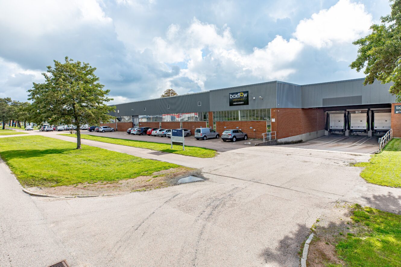 En stor industrihall med lastkajer, parkerade bilar och en omgivande gräsyta under en delvis molnig himmel.