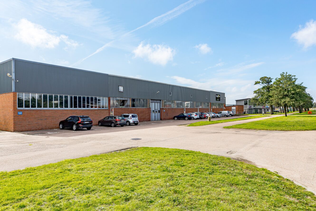 Ett stort industriellt lager med en nedre fasad i tegel och gråa övre väggar, flera parkerade bilar utanför och en gräsyta i förgrunden under en blå himmel.