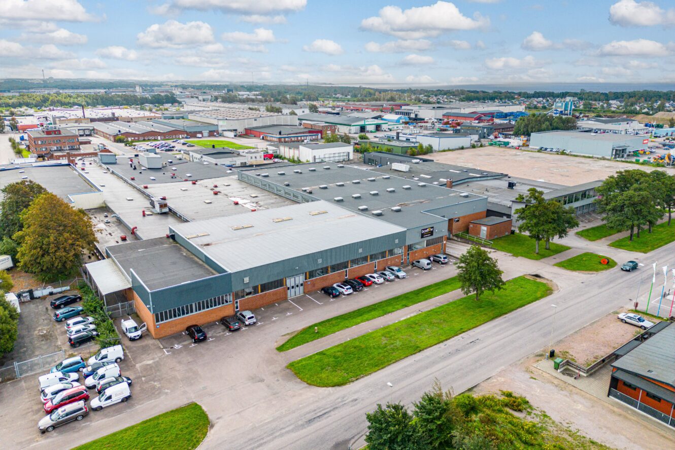 Flygfoto över ett stort industrikomplex med flera lagerlokaler, parkeringsplatser, vägar och omgivande byggnader under en delvis molnig himmel.