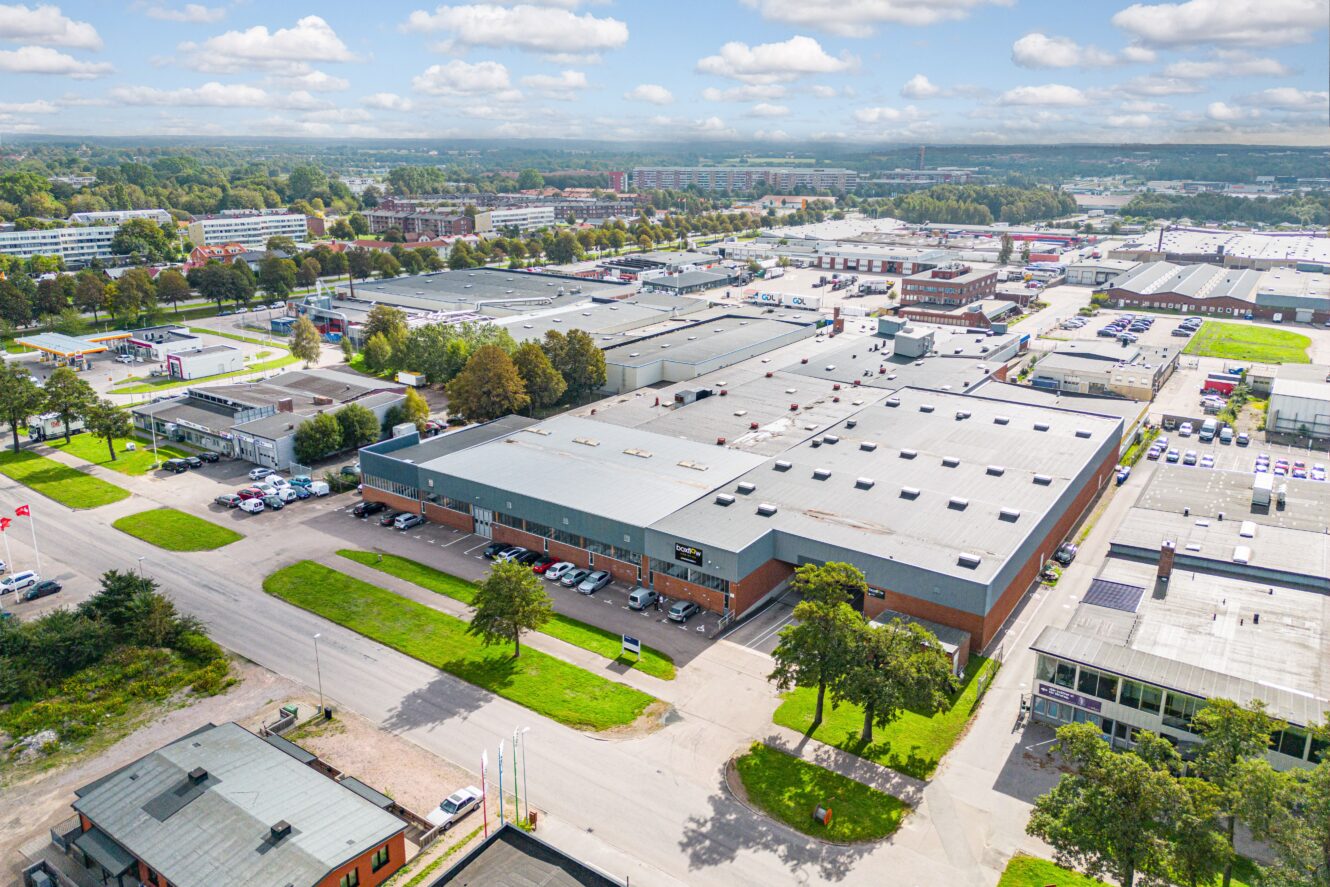 Flygfoto över en industripark med stora lagerbyggnader, parkeringsplatser, träd och omgivande vägar under en delvis molnig himmel.