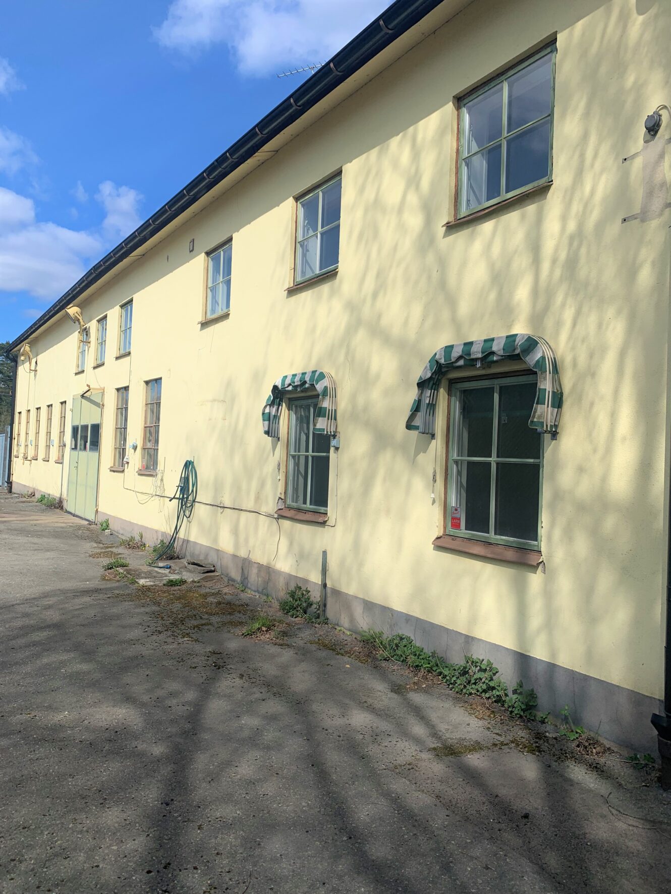 En lång, gul tvåvåningsbyggnad med flera fönster, varav två har grönvitrandiga markiser, mot en blå himmel med några moln.