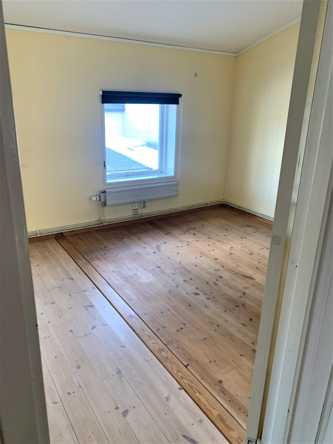 Tomt rum med ljust trägolv, beige väggar, ett enda fönster med svart persienn och en vit radiator under fönstret.
