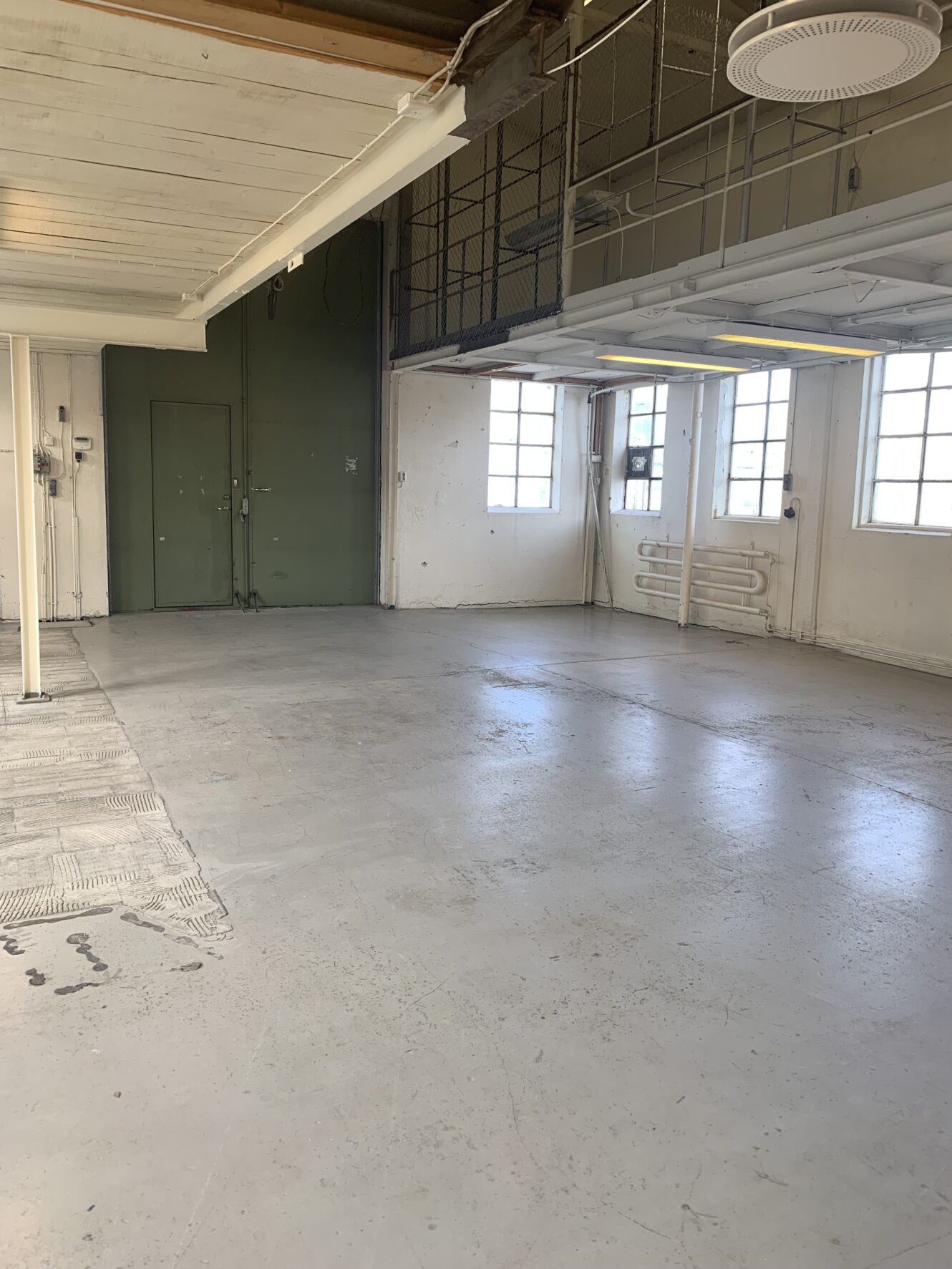 Tomt industrirum med betonggolv, vita väggar, stora fönster, synliga rör och en grön dubbeldörr. Takbelysning och delvis heltäckningsmatta är synliga.