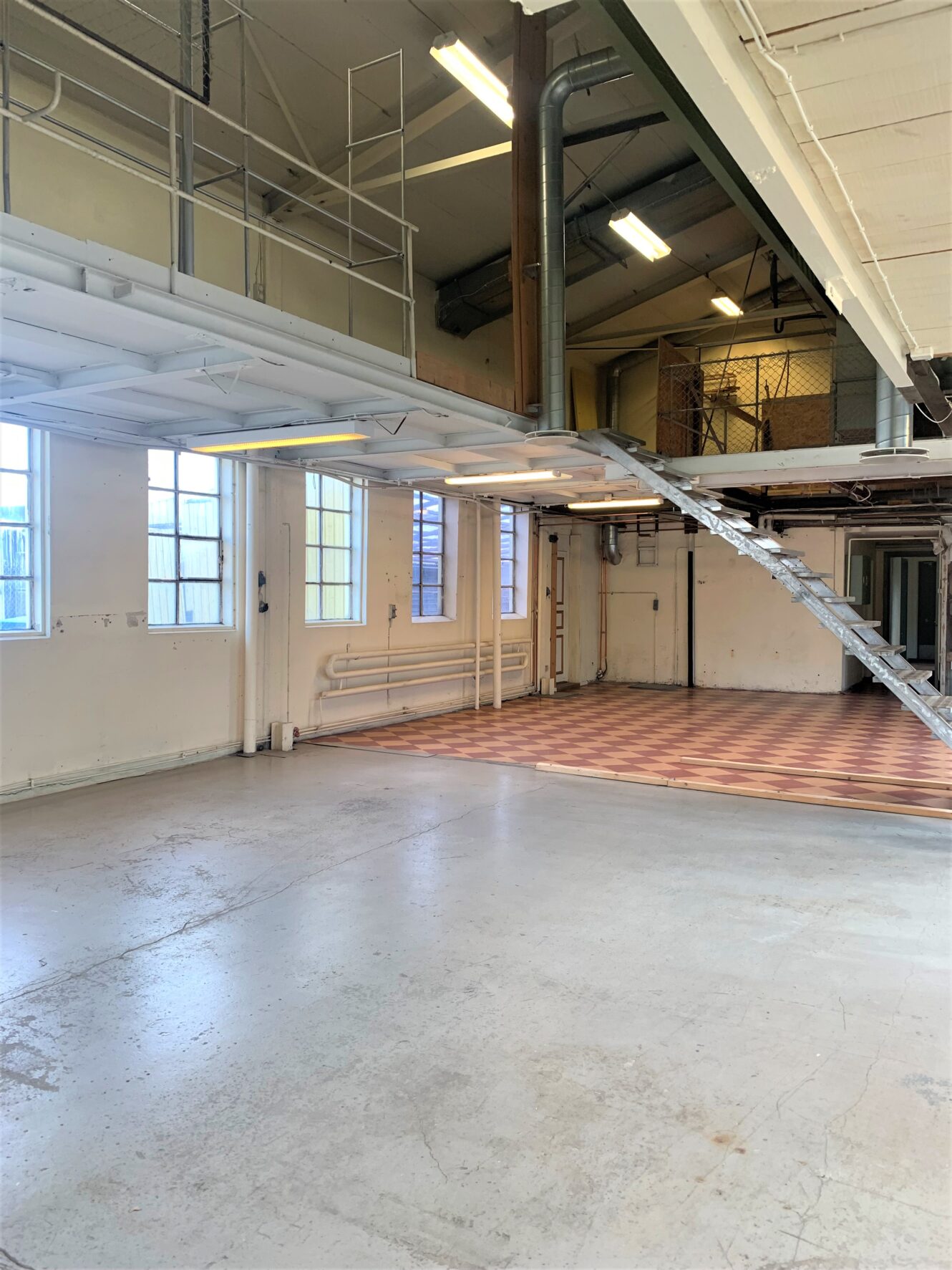 Inredning i industriell stil med högt i tak, rutigt klinkergolv, metalltrappa som leder till mezzanin och stora fönster med naturligt ljusinsläpp.