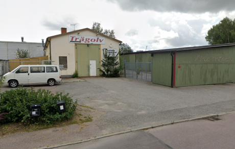 En liten industribyggnad med skylten Trägolv och en vit skåpbil parkerad utanför, bredvid en grön metallklädd byggnad en molnig dag.