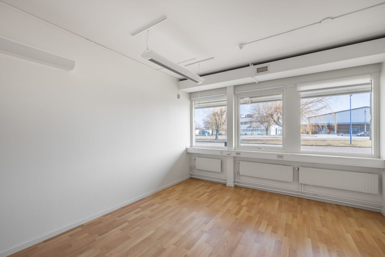 Tomt rum med vita väggar, ljust trägolv och stora fönster som släpper in naturligt ljus. Radiatorer löper under fönstren och taklampor är installerade ovanför.
