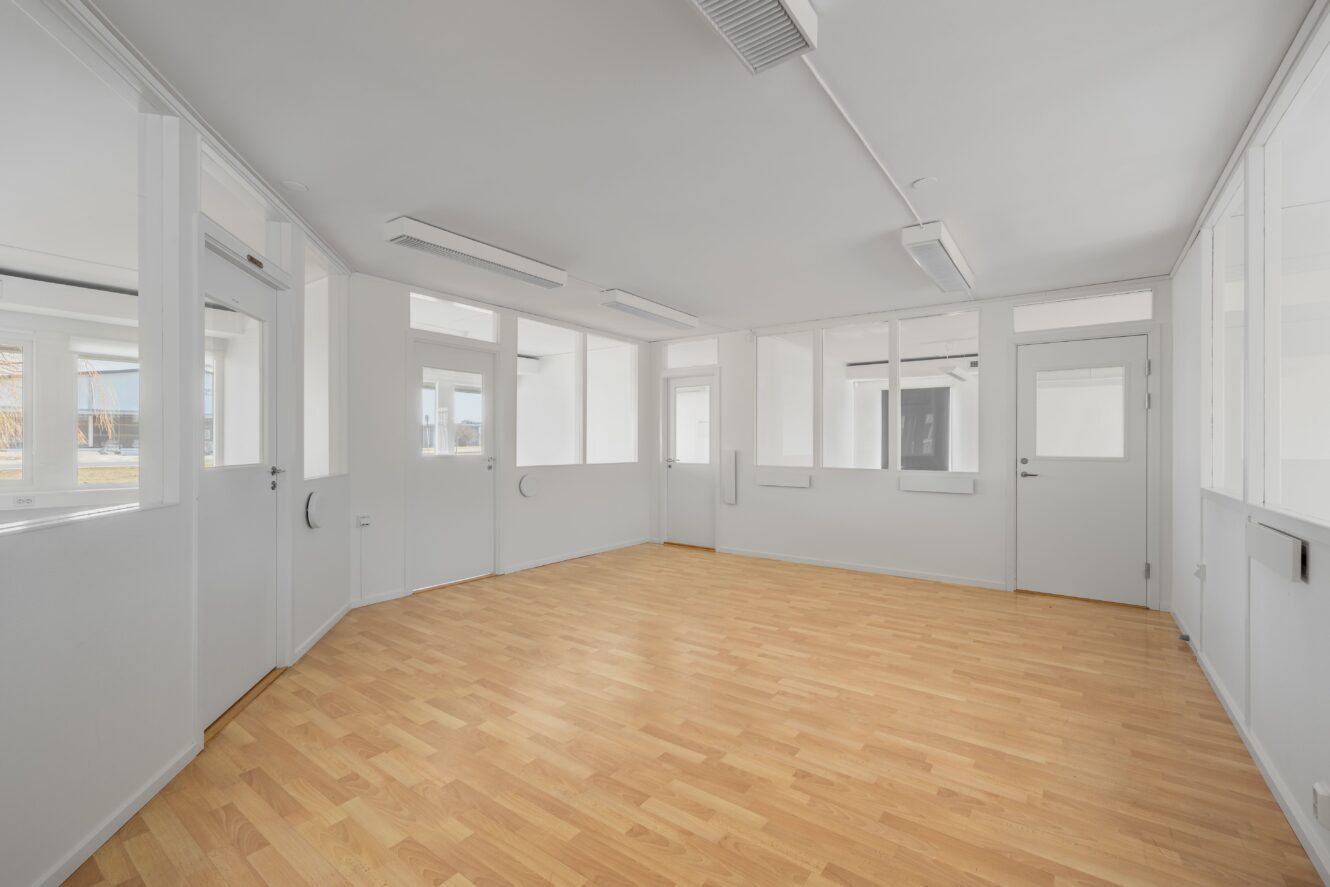 Tomt rum med ljust trägolv, vita väggar, flera dörrar och stora fönster som släpper in naturligt ljus.