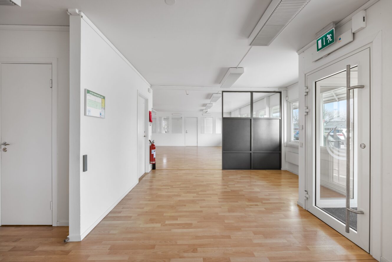 En modern, tom kontorskorridor med ljust trägolv, vita väggar, stora fönster, en glasdörr och en svart skiljevägg.