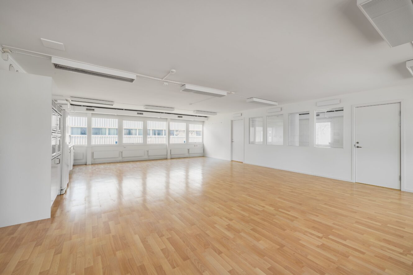Tomt, rymligt rum med ljust trägolv, vita väggar, flera fönster på ena sidan och en dörr på den högra. I taket finns flera armaturer och ventilationsöppningar.