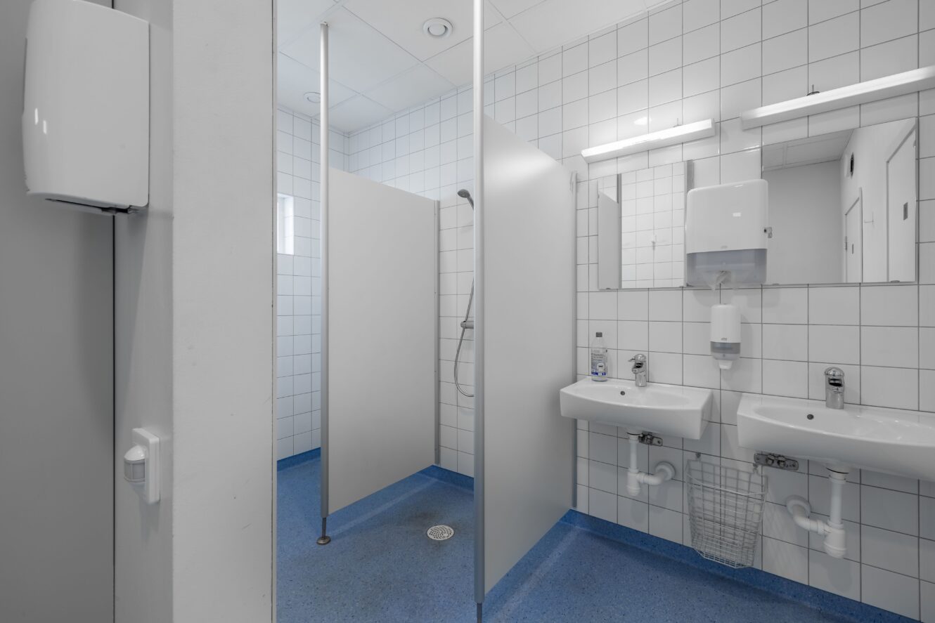 En offentlig toalett med vitkaklade väggar, blått golv, två handfat med speglar, tvålbehållare och ett duschutrymme som är avskilt med skiljeväggar.