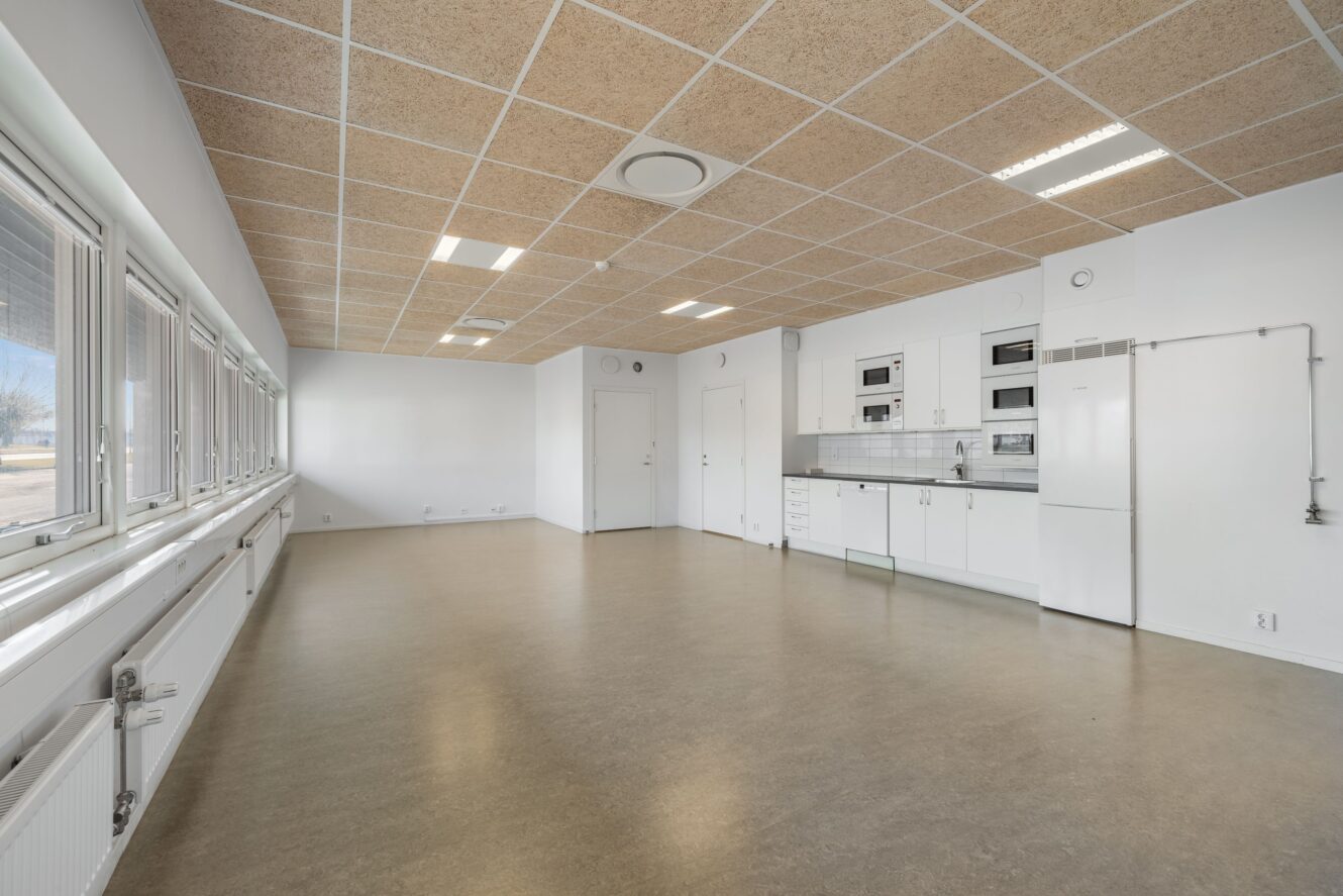 Rymligt, tomt rum med stora fönster, vita väggar, beige tak och ett pentry med vita skåp och vitvaror på höger sida.