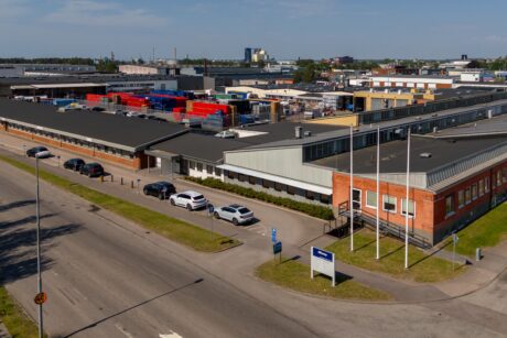 Flygfoto över ett industrikomplex med lagerbyggnader, parkerade bilar, containrar och en gata i förgrunden.