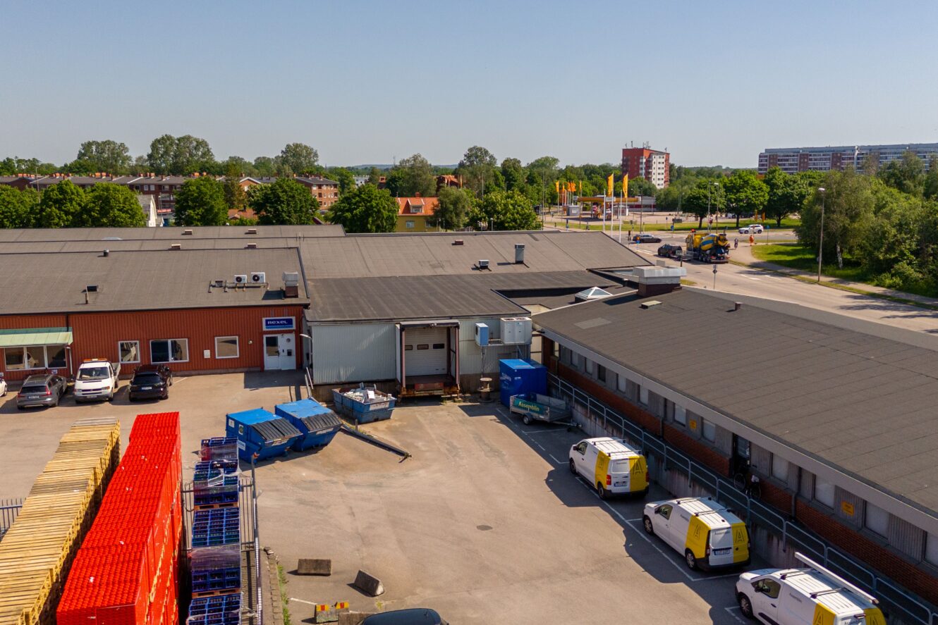 Ett lager med lastkajer, flera skåpbilar, staplade pallar och containrar på en asfalterad tomt; träd och flerfamiljshus syns i bakgrunden.