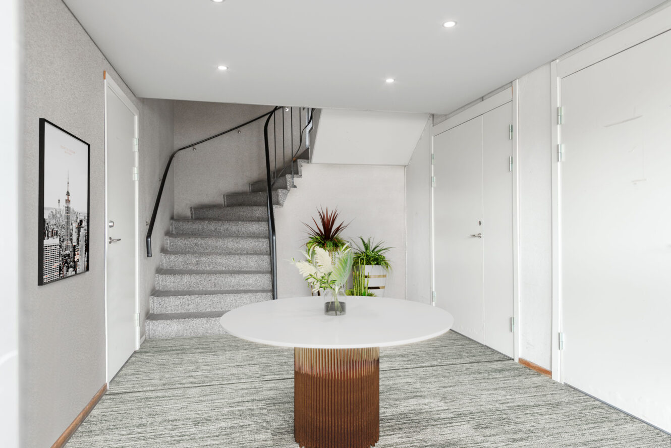 En modern entré med ett runt vitt bord, krukväxter, en trappa med svart räcke och vita dörrar; en inramad tavla hänger på den vänstra väggen.