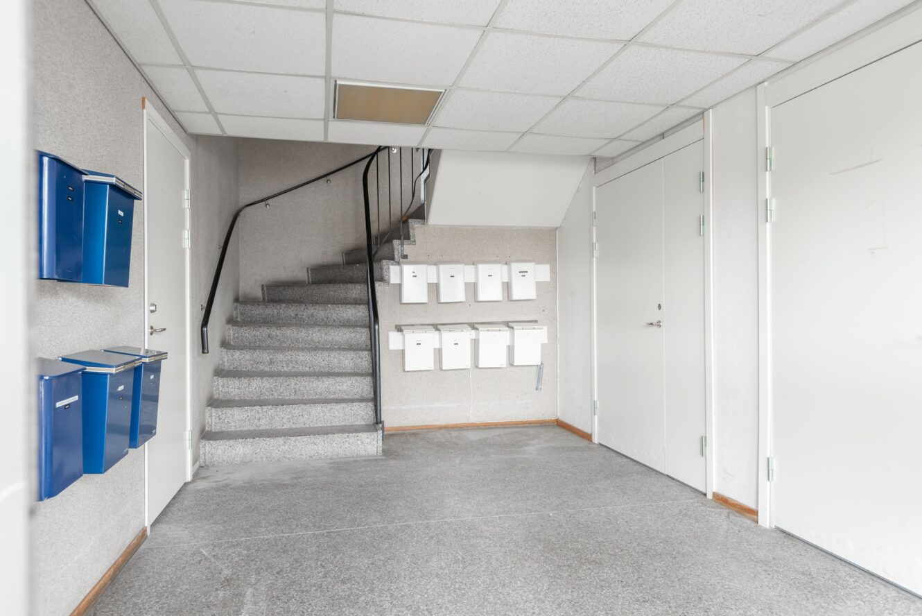 En ren entré till ett hyreshus med grå trappor, blå och vita brevlådor på väggarna, vita dörrar och ett kaklat tak.