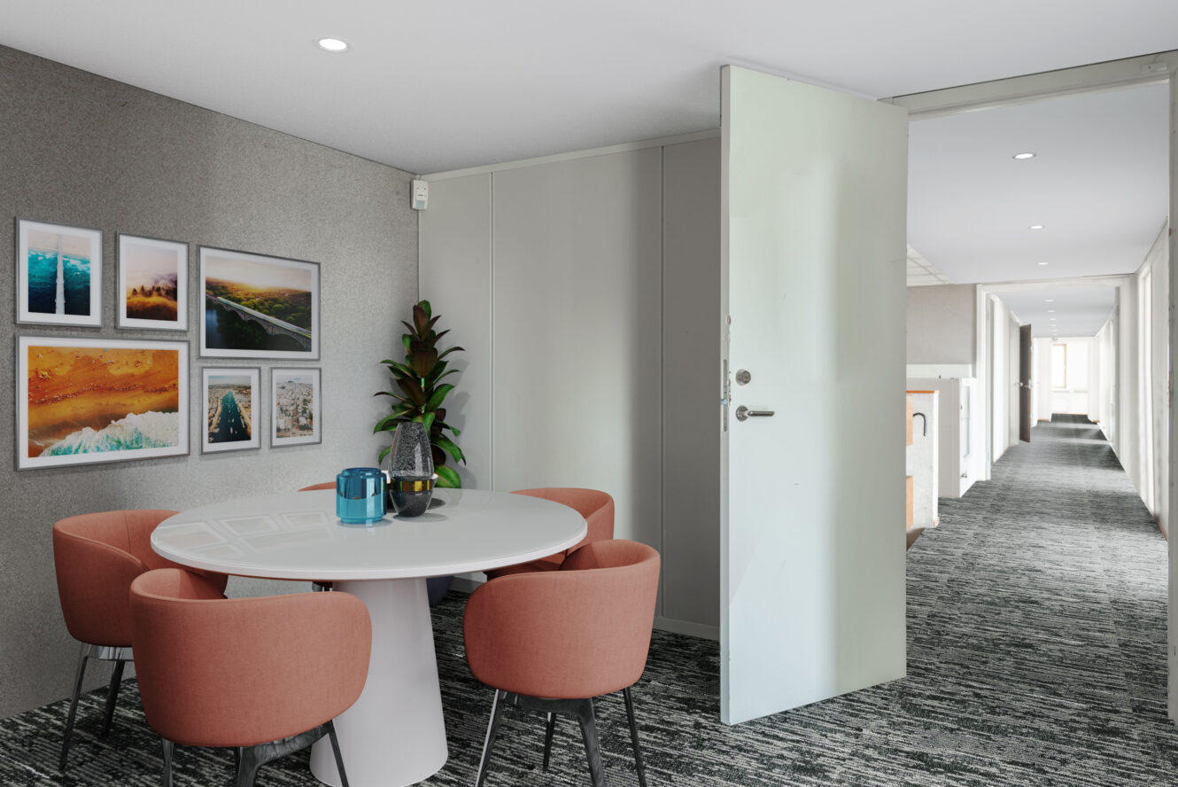 Ett runt vitt bord med fyra rosa stolar står vid en vägg med inramade foton och en krukväxt, bredvid en öppen dörr som leder till en lång korridor.